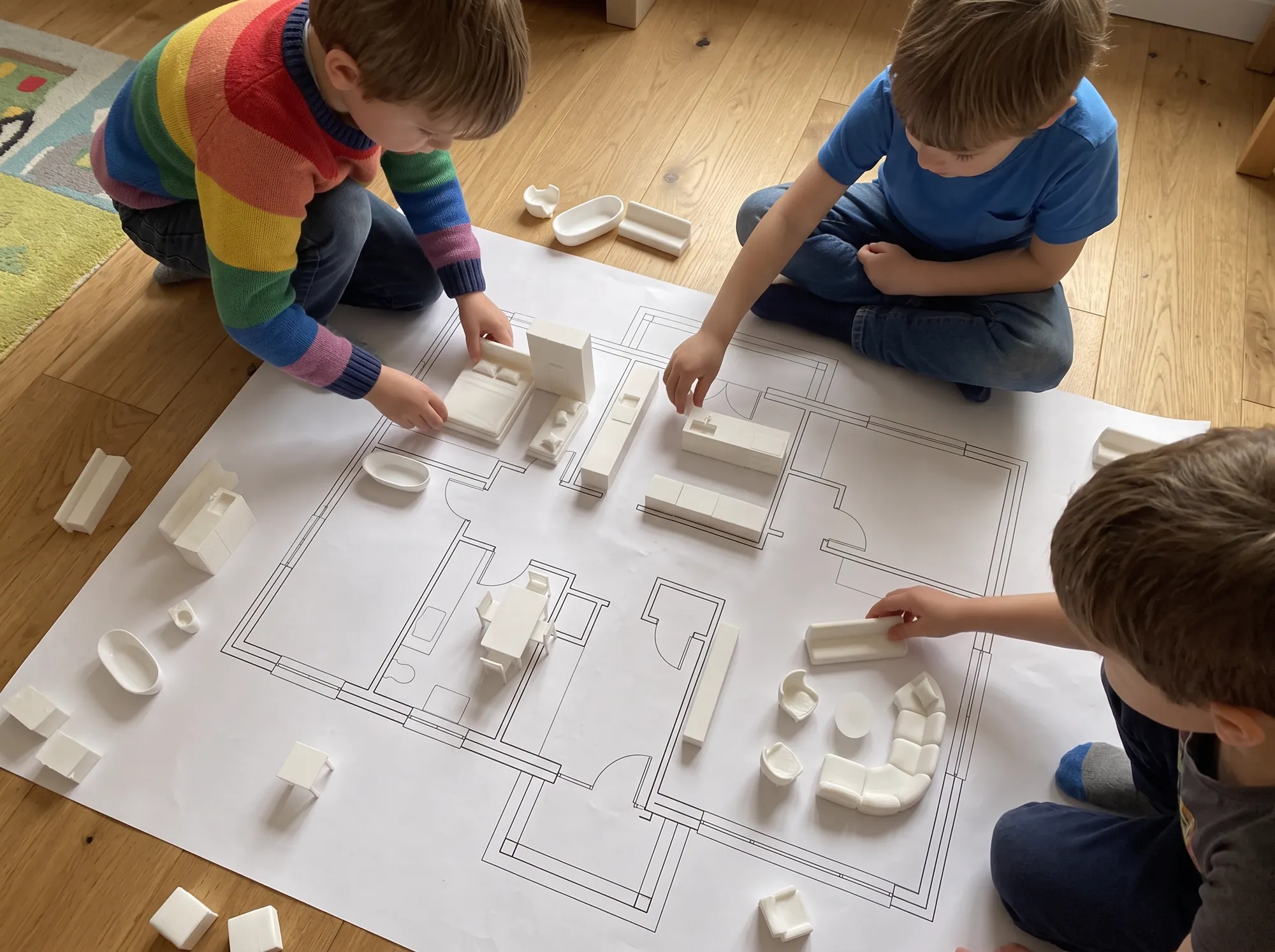 Troje dzieci na podłodze układa meble z drukarki 3D na dużym planie mieszkania wydrukowanym w skali
